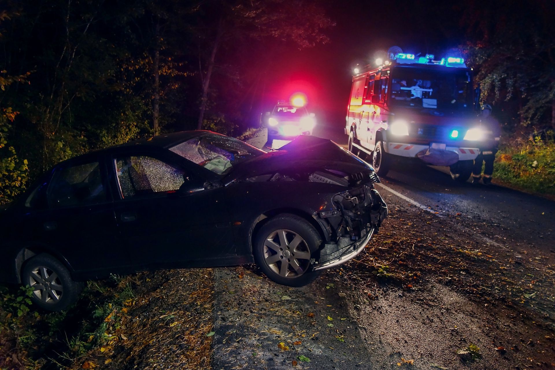 Une voiture noire accidentée de nuit, à côté de véhicules de police et d'urgence dont les gyrophares sont allumés, sur une route sombre.