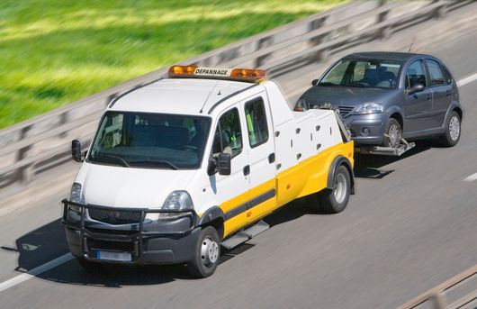 Une dépanneuse remorque une voiture grise sur une autoroute bordée d'herbe et de glissières de sécurité.