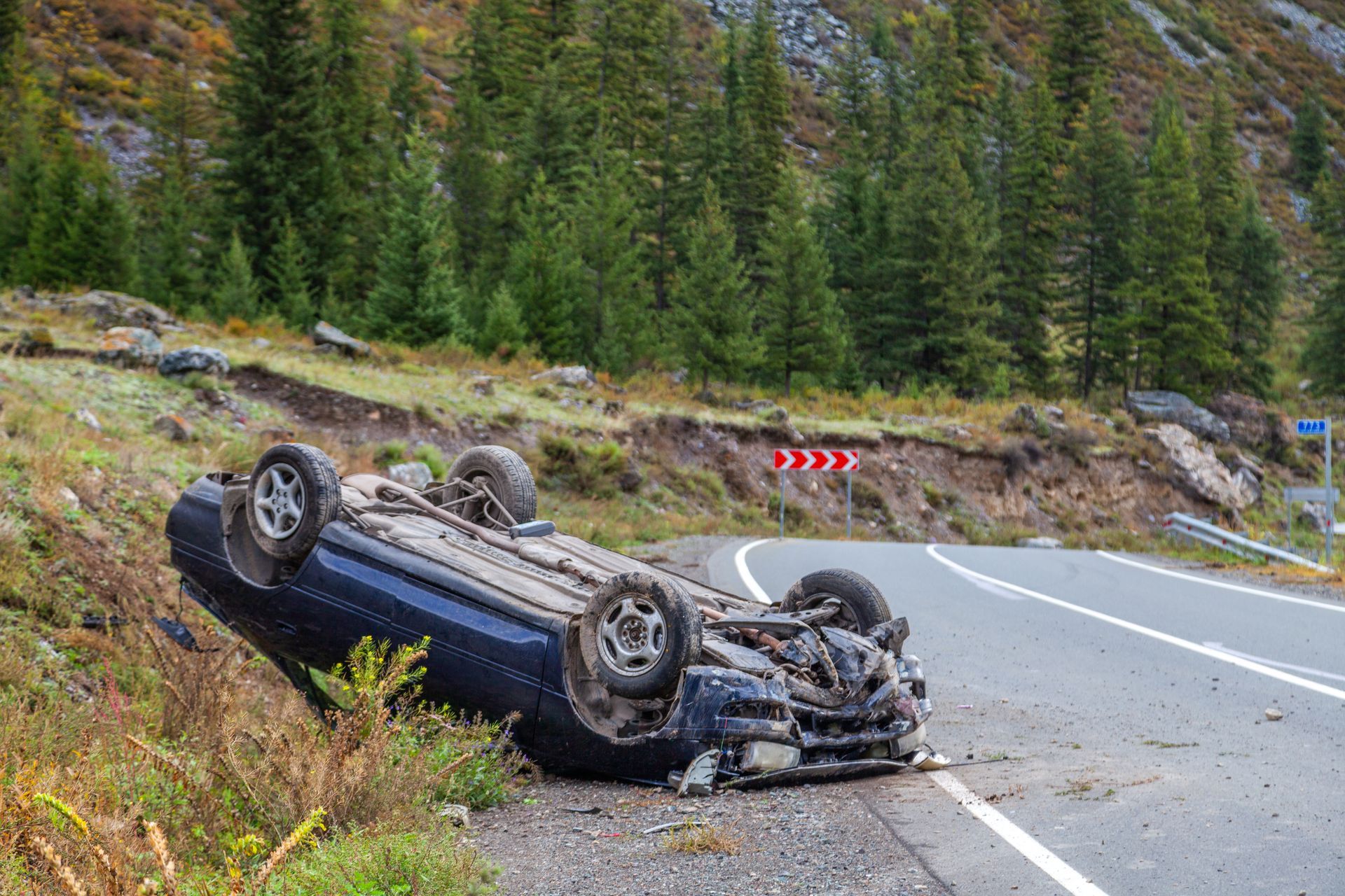 Voiture bleue renversée au bord d'une route de montagne, près d'une route sinueuse et d'un versant boisé.