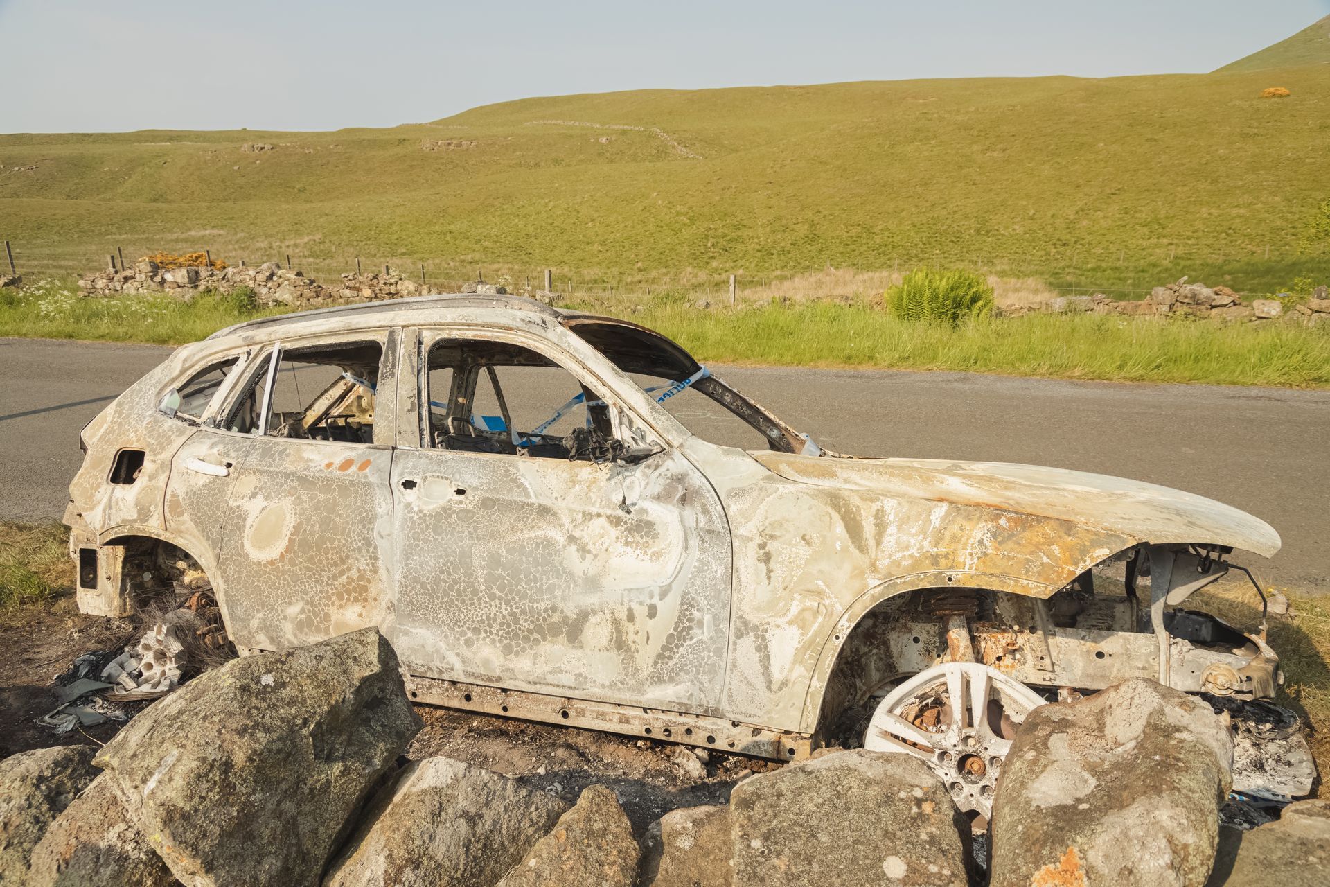 Une épave de SUV blanc calcinée gît sur des rochers au bord d'une route dans un paysage rural herbeux.