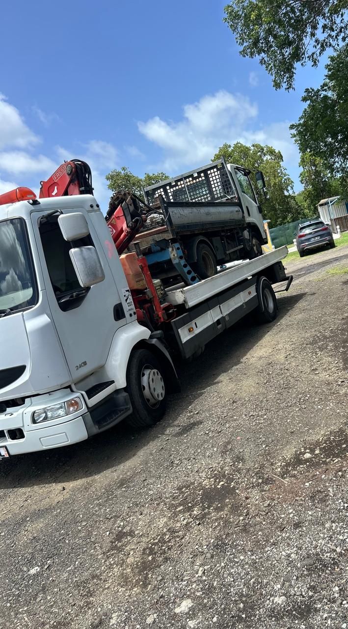 Une dépanneuse blanche à plateau transportant un petit camion noir sur le bord d'une route de gravier sous un ciel bleu