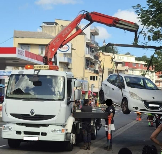 Une dépanneuse soulève une voiture blanche dans une rue de la ville, près d'immeubles d'habitation.