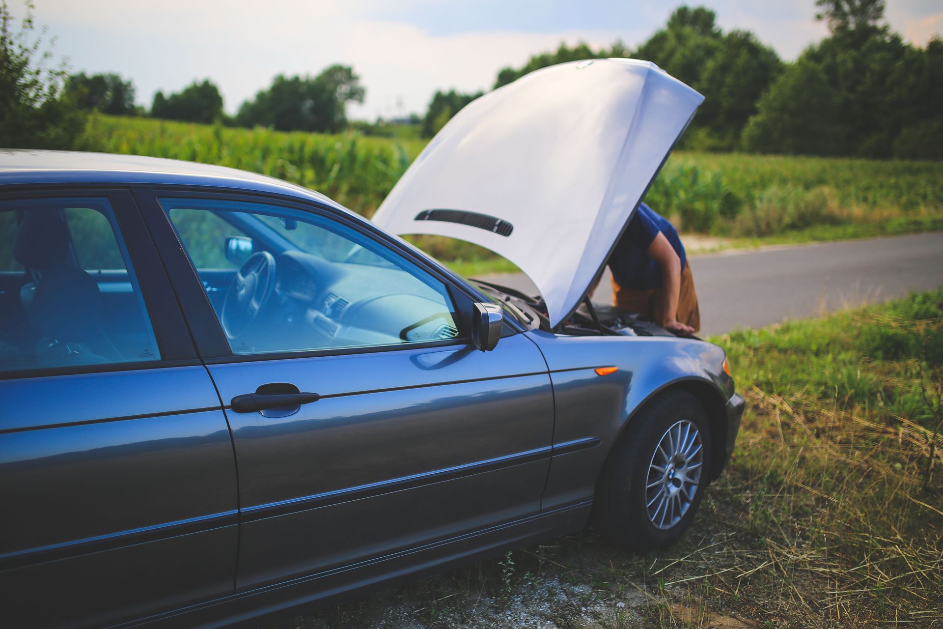 Voiture bleue, capot ouvert, garée en bord de route, laissant apparaître une personne inspectant le moteur à l'avant.