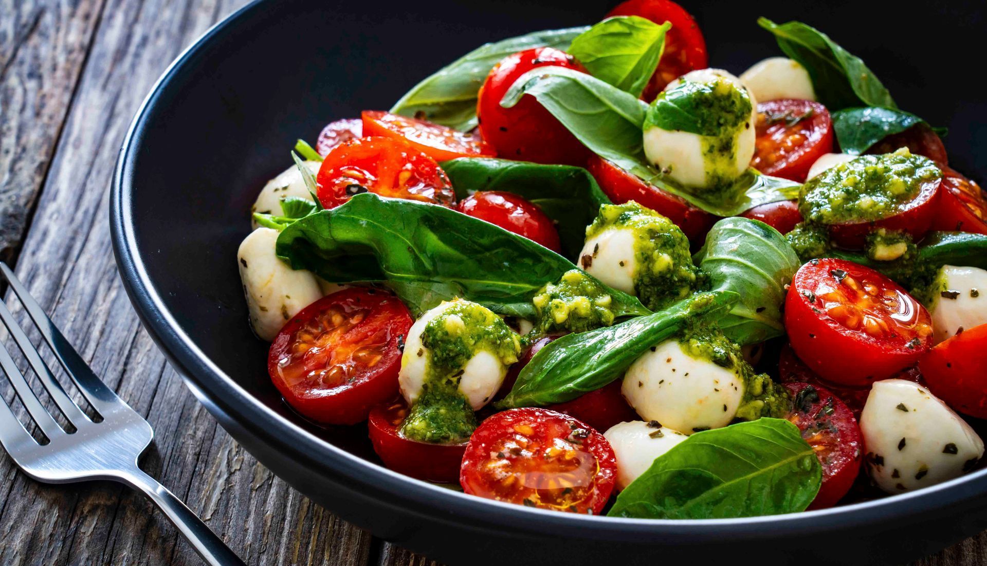 Ein Salat mit Tomaten, Mozzarella, Basilikum und Pesto in einer Schüssel mit einer Gabel.