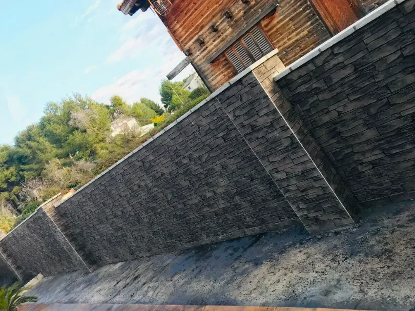 Vista inclinada de un muro de piedra oscura junto a un edificio de madera, con árboles y cielo azul al fondo.
