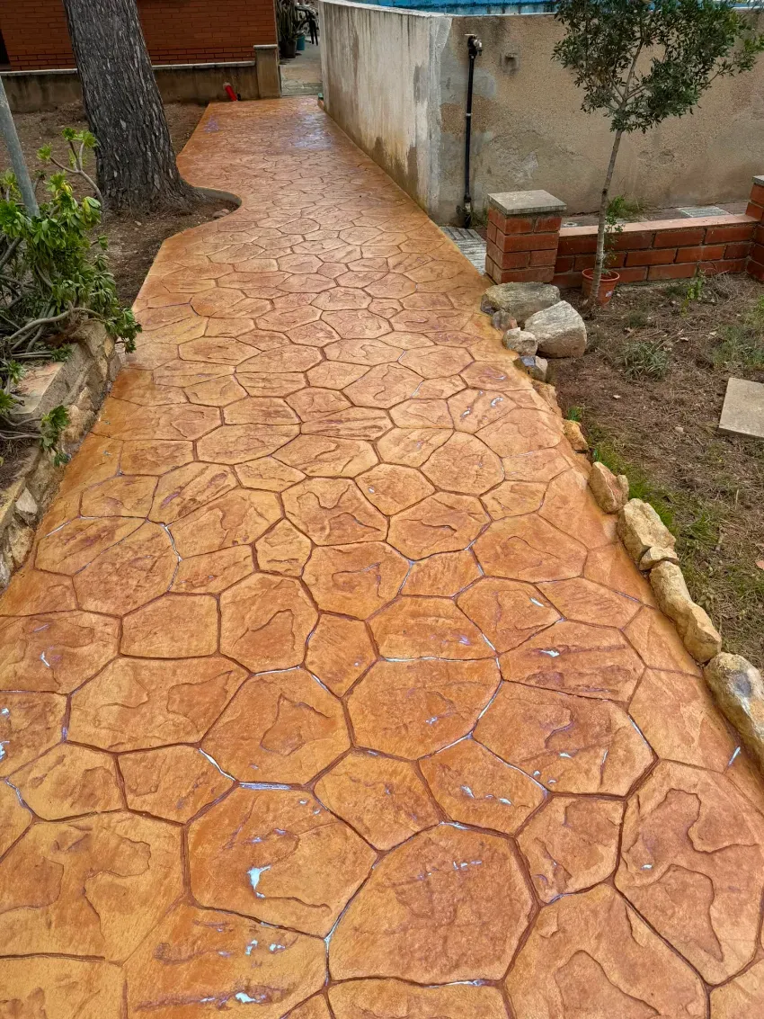 Sendero con estampado naranja junto a una casa y un jardín, con un árbol a la izquierda.