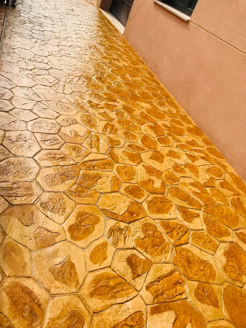 Un sendero de color naranja con diseño de piedra, junto a una pared y una ventana de color melocotón.