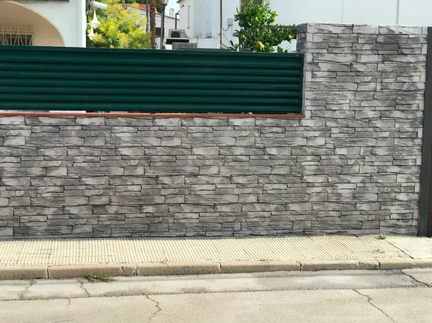 Muro de piedra gris con una valla verde y un bordillo a lo largo de una acera al aire libre.