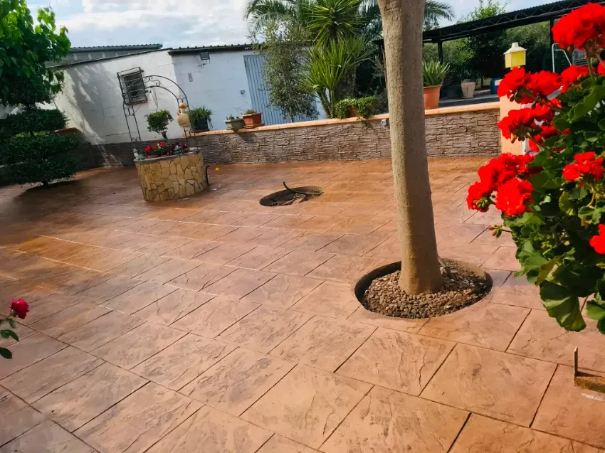 Patio embaldosado y empapado por la lluvia con un árbol, una jardinera de piedra y flores rojas junto a una pared blanca.