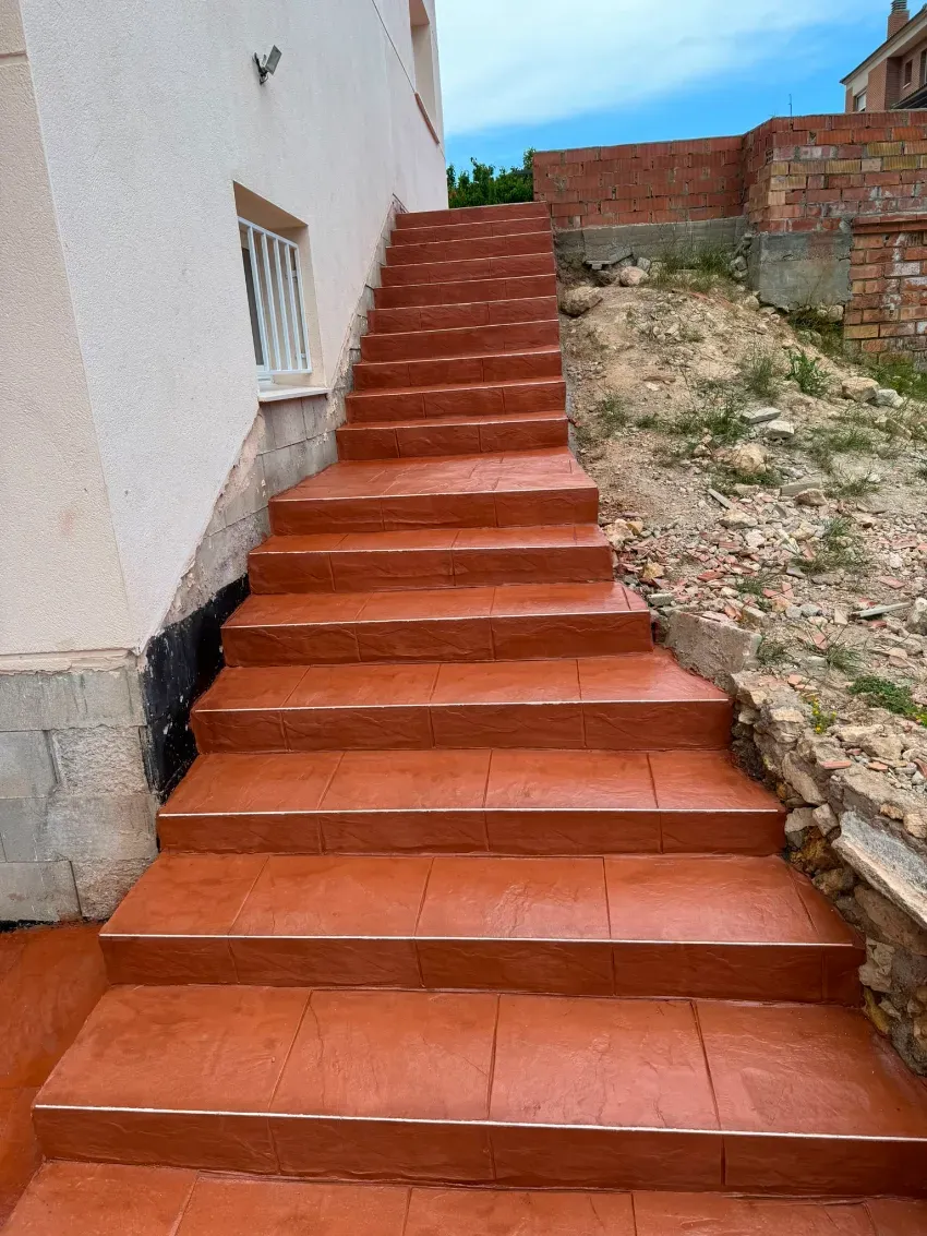 Una estrecha escalera de terracota junto a una pared blanca y una ladera rocosa bajo un cielo azul.