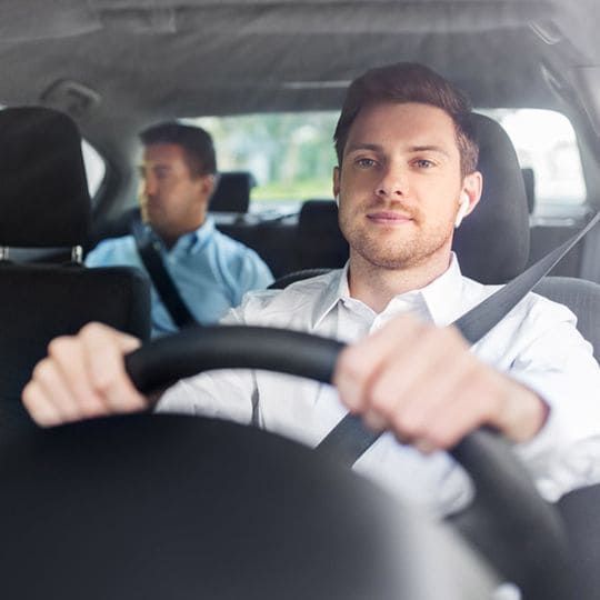 Una persona conduce un coche con un pasajero en el asiento trasero; ambos visten camisas blancas y azules y llevan puesto el cinturón de seguridad.
