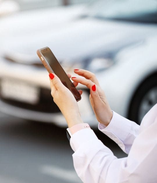 Persona con las uñas pintadas de rojo usando un teléfono inteligente al aire libre, con un coche borroso al fondo.