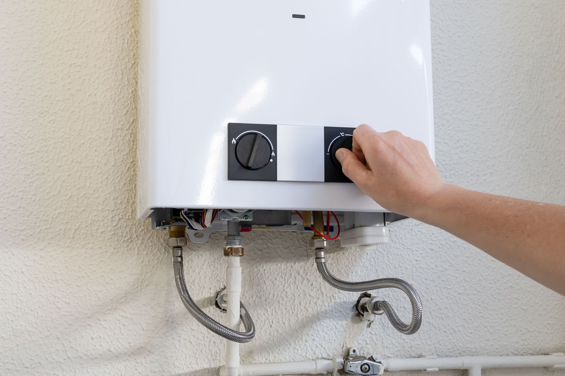 Main ajustant le bouton de commande d’un chauffe-eau blanc fixé au mur, avec des tuyaux apparents.
