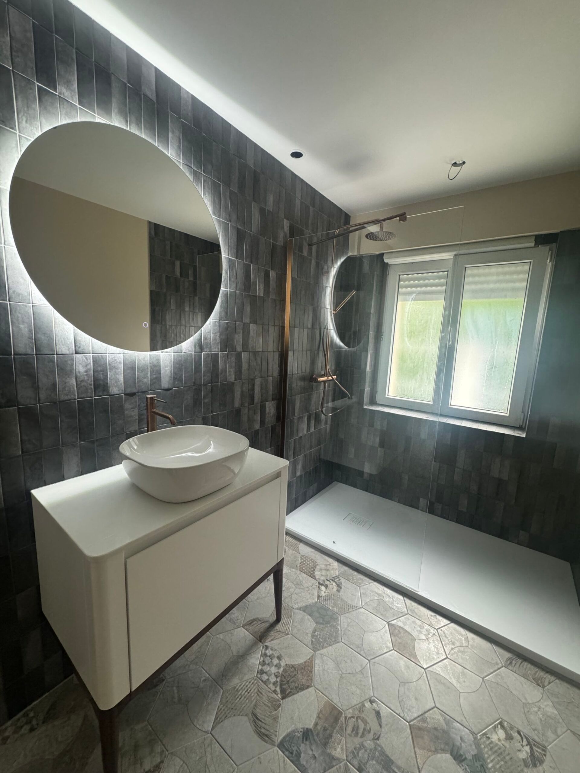 Salle de bains moderne avec miroir rond, meuble-lavabo blanc, murs en carrelage foncé et douche à l’italienne.