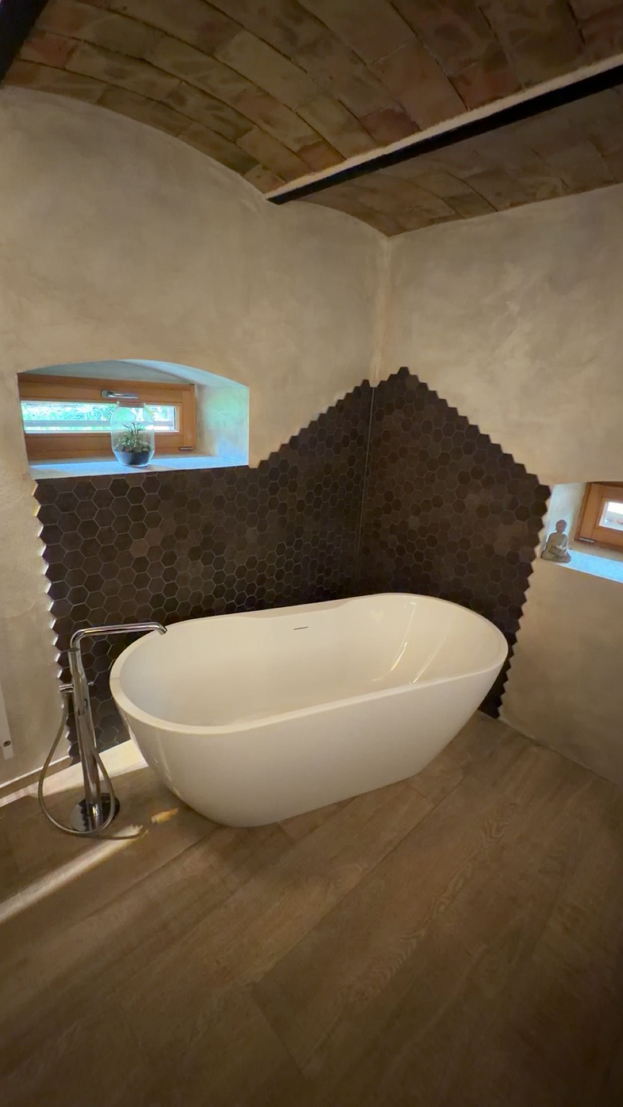 Salle de bains moderne avec baignoire blanche îlot, mur carrelé foncé, parquet en bois et petites fenêtres