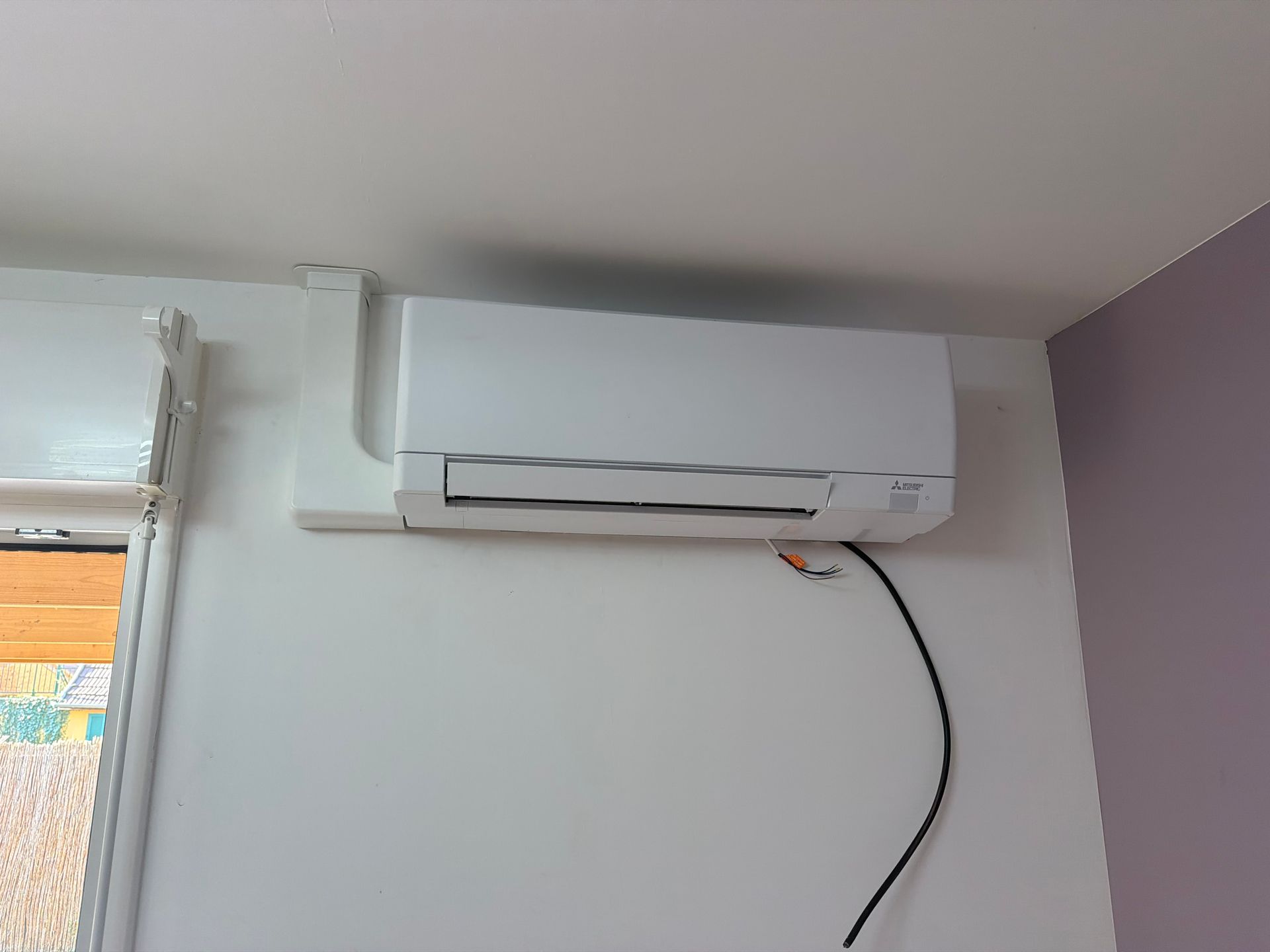 Climatiseur mural blanc au-dessus d’une fenêtre, avec un cordon d’alimentation noir qui pend.
