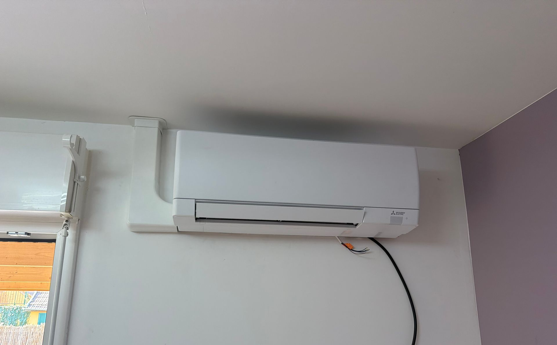 Climatiseur blanc mural au-dessus d’une fenêtre, avec un câble d’alimentation qui pend contre un mur clair.