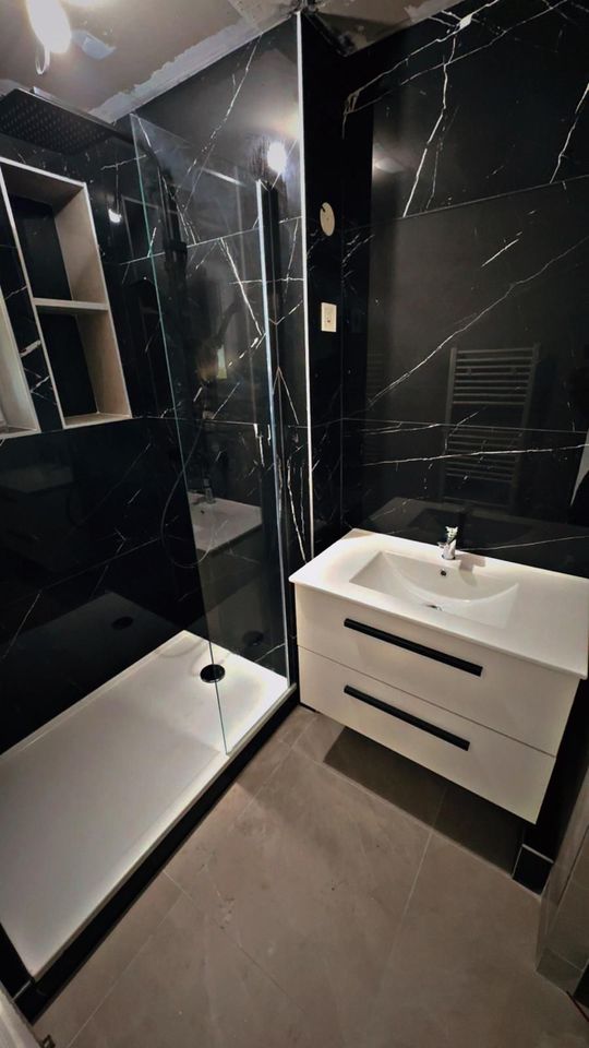 Salle de bains moderne et compacte, avec des murs en marbre noir, une douche en verre et un meuble vasque blanc