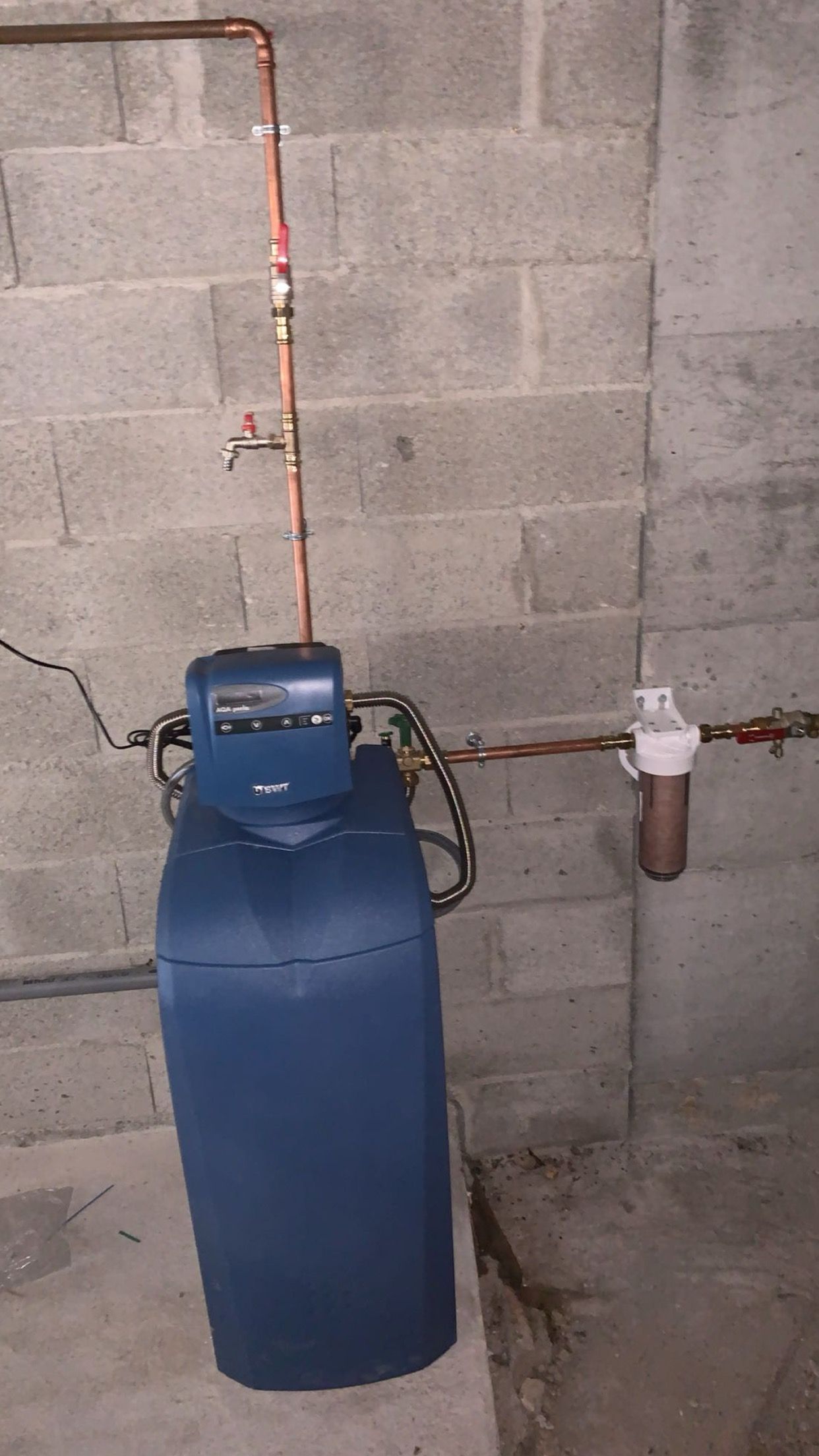 Réservoir bleu d’adoucisseur d’eau avec des tuyaux en cuivre, contre un mur en parpaings dans une buanderie