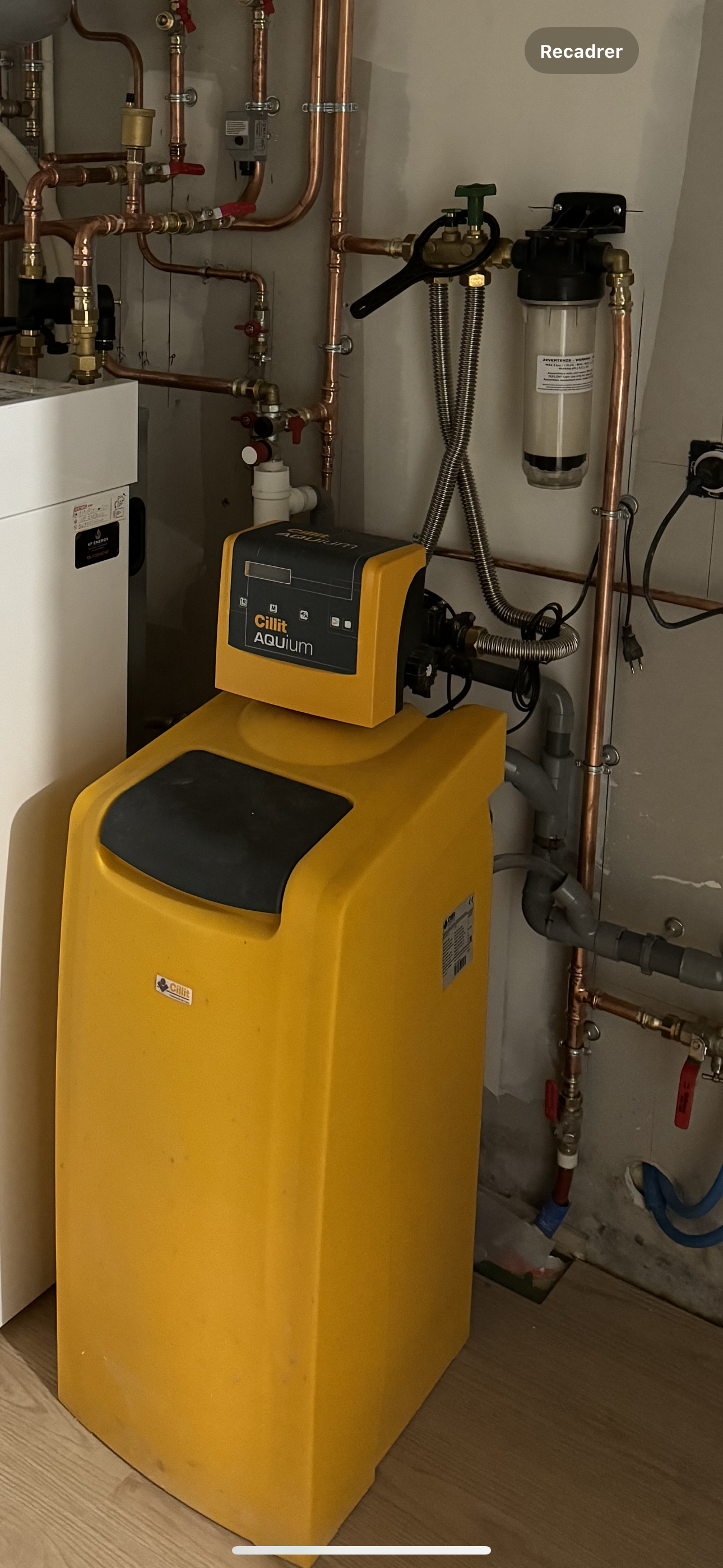 Réservoir jaune d’adoucisseur d’eau dans un sous-sol, à côté de tuyaux en cuivre, d’une chaudière et d’un système de filtration.