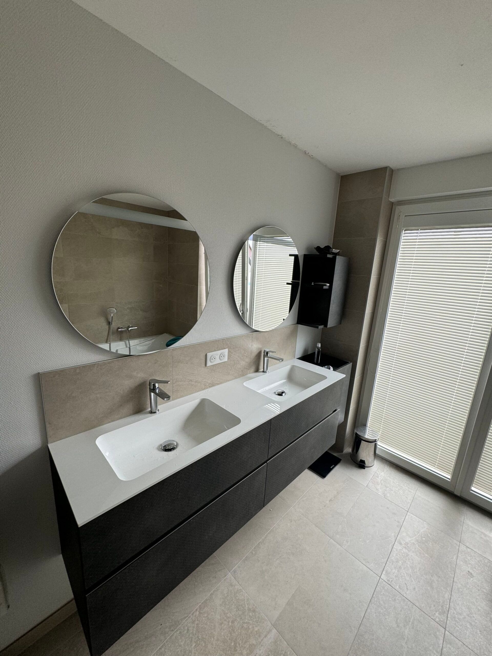 Meuble-lavabo moderne avec double vasque, miroir rond et stores à la fenêtre près de la douche.