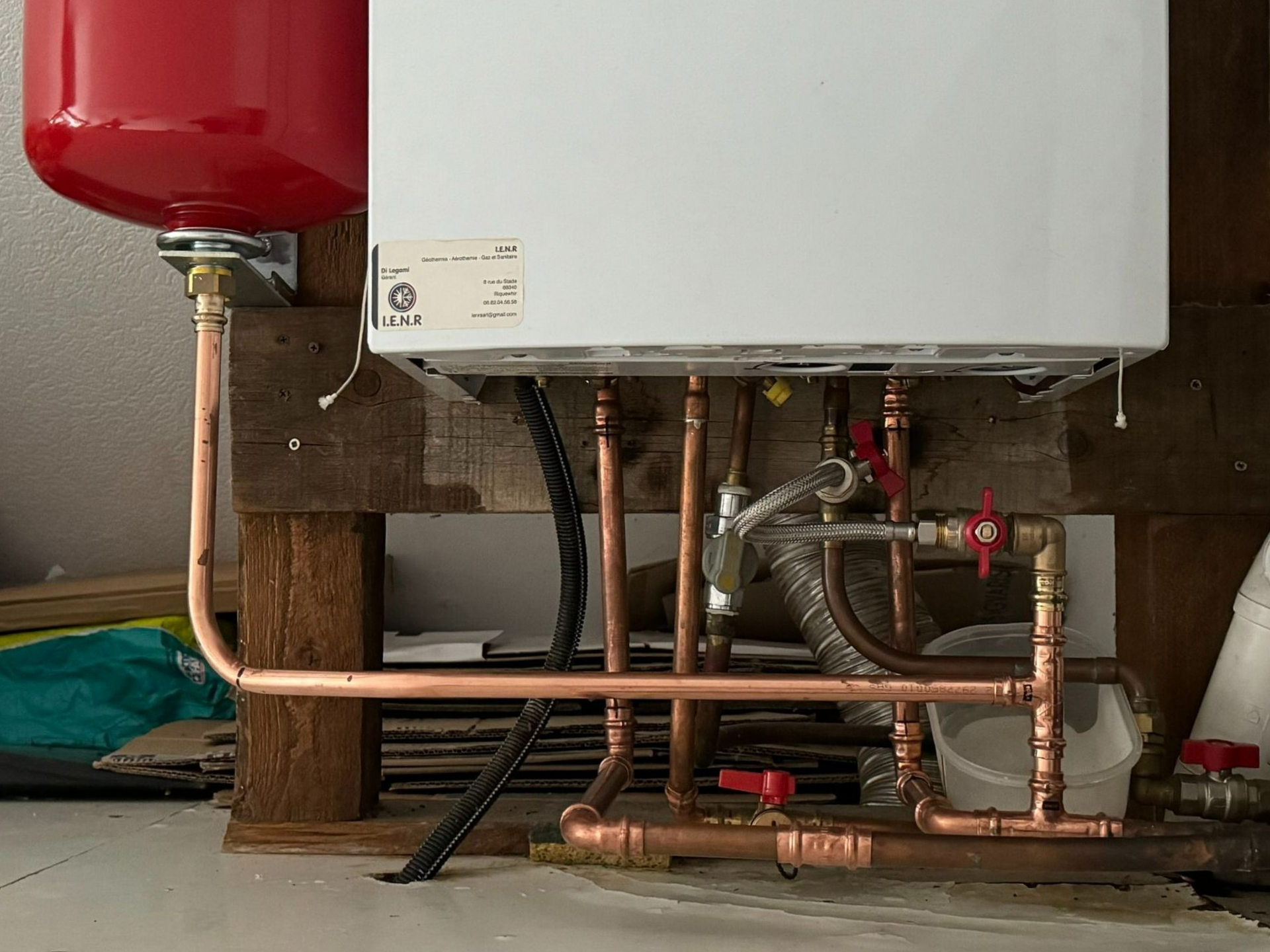 Plomberie d’un chauffe-eau avec un vase d’expansion rouge et des tuyaux en cuivre sous un réservoir fixé au mur.