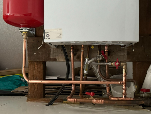 Plomberie d’un chauffe-eau avec un vase d’expansion rouge et des tuyaux en cuivre sous un réservoir fixé au mur.