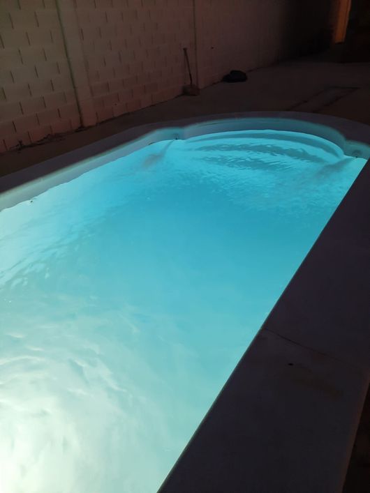 Piscina iluminada que brilla en azul por la noche junto a una terraza oscura y una pared de ladrillo.