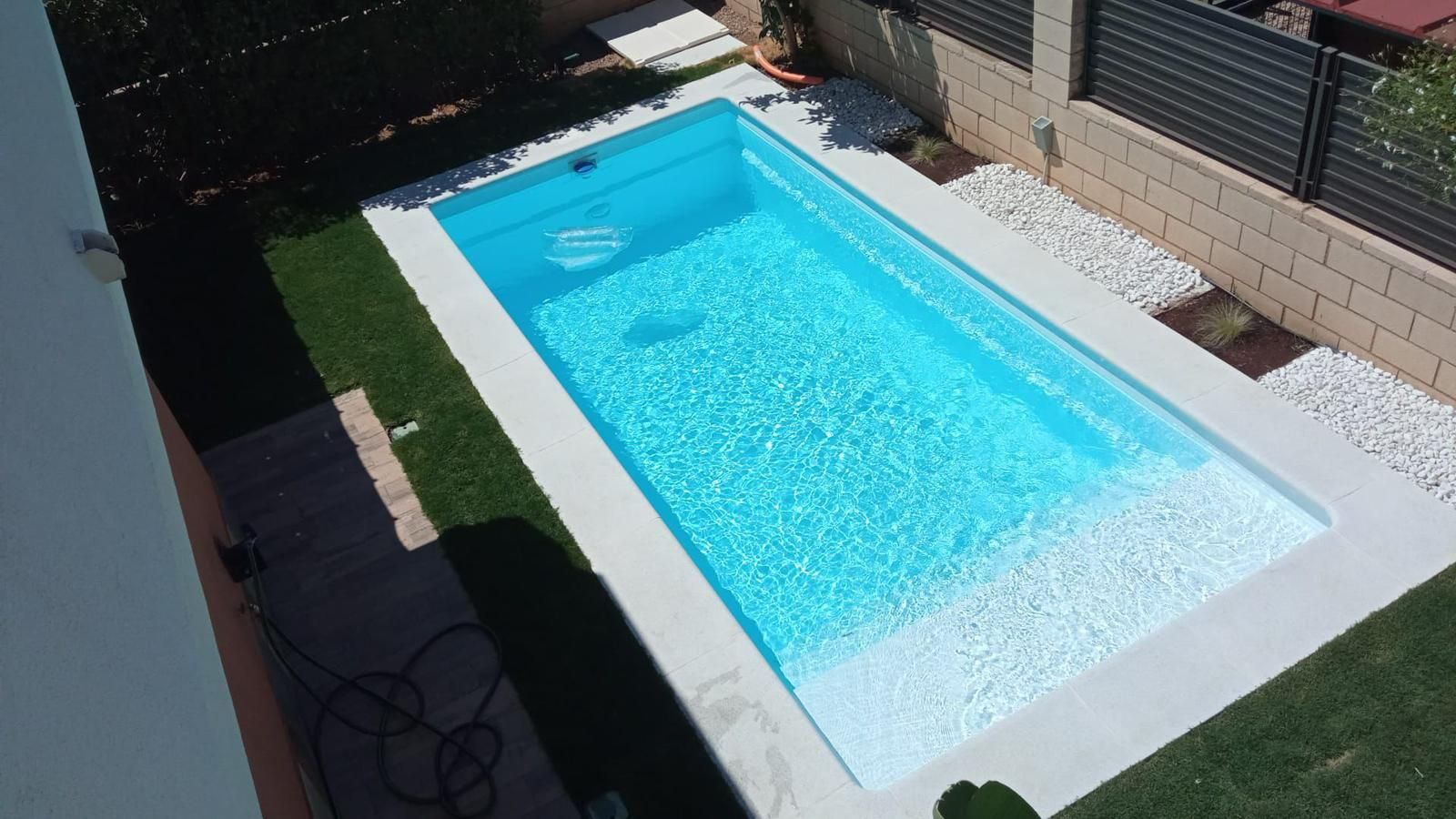 Piscina rectangular en el patio trasero con agua azul brillante, borde blanco y terraza de guijarros bañada por el sol.