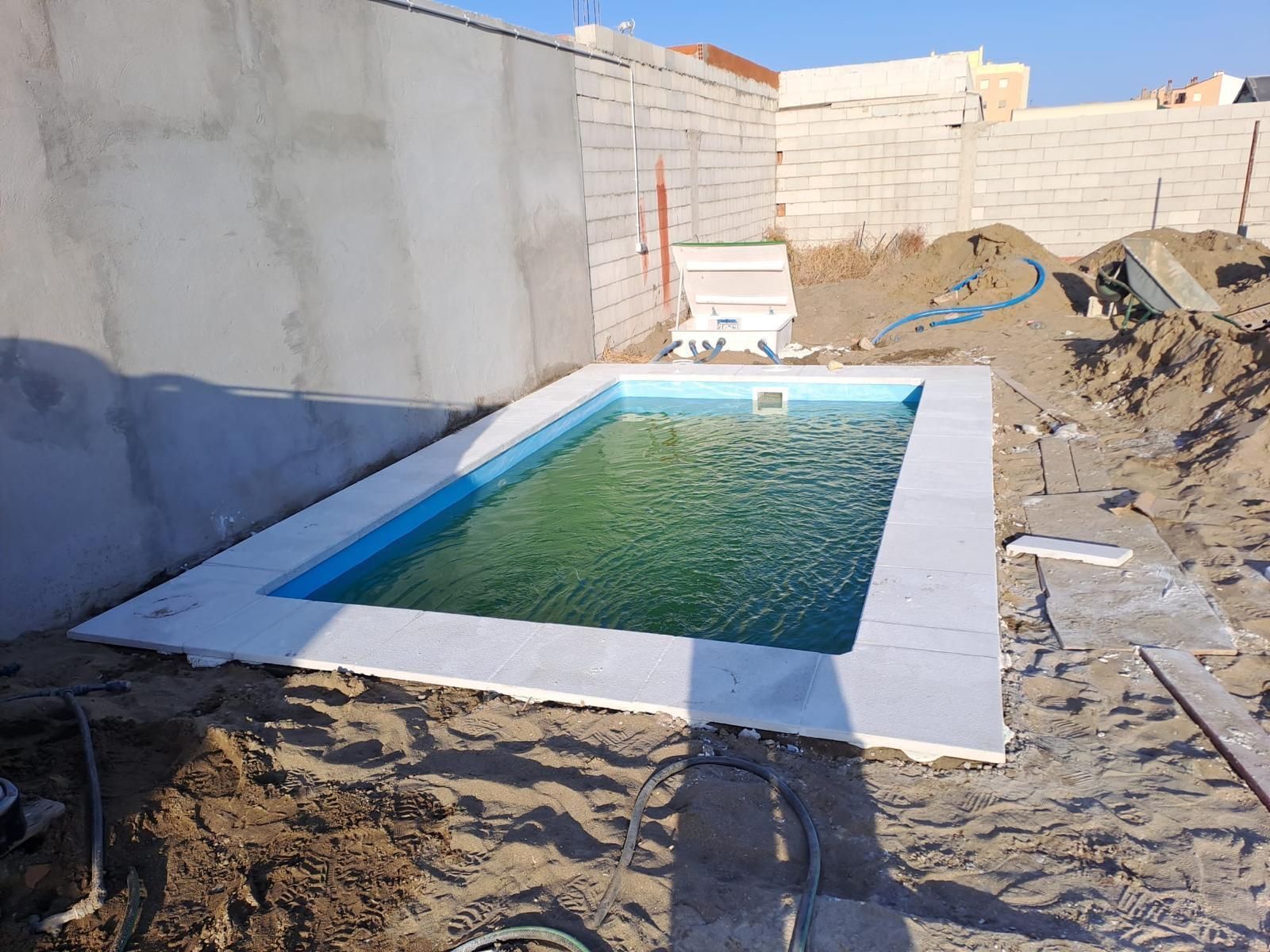 Pequeña piscina rectangular en el patio trasero, rodeada de arena y paredes sin terminar, con agua verde.