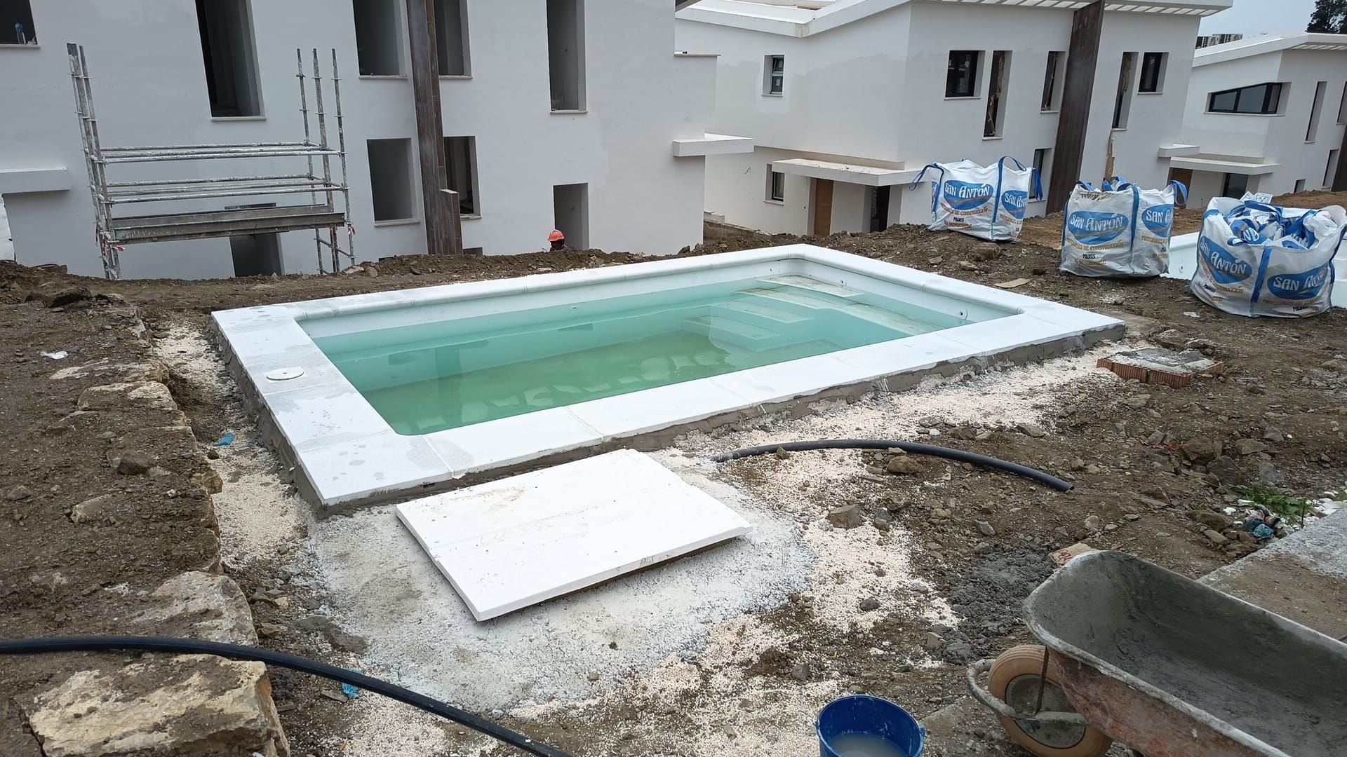 Piscina rectangular en construcción en el patio trasero, con borde blanco y tierra y materiales de construcción a su alrededor.