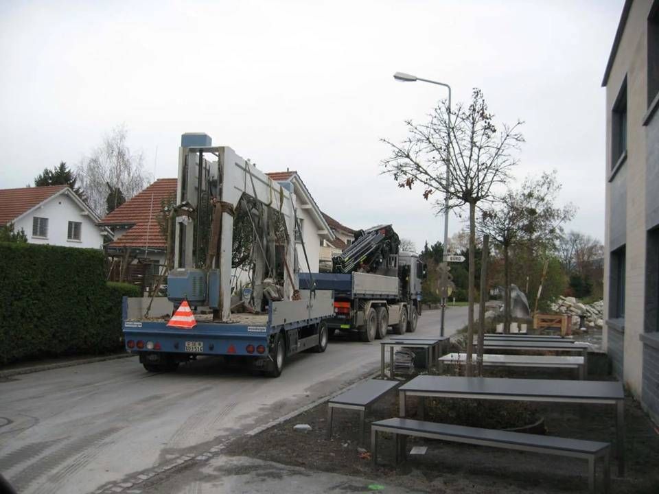 Ein Lastwagen transportiert eine grosse Metallkonstruktion durch eine Wohnstrasse mit Häusern und Bäumen in der Nähe.