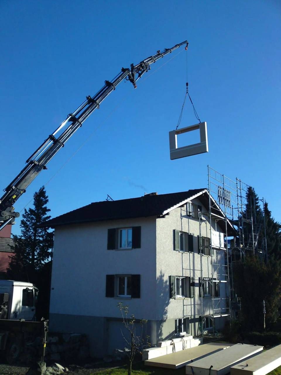 Ein Kran hebt an einem klaren Tag einen grossen Fensterrahmen über einem weissen Wohnhaus an.