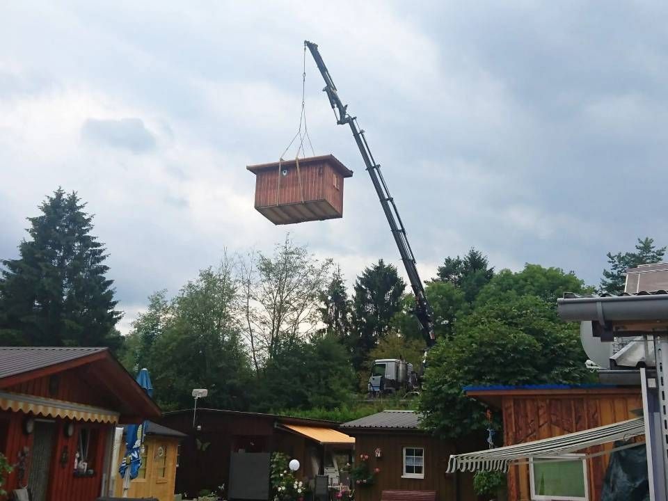 Ein Kran hebt ein Holzkabinensegment über Bäume in einem Hof unter bewölktem Himmel