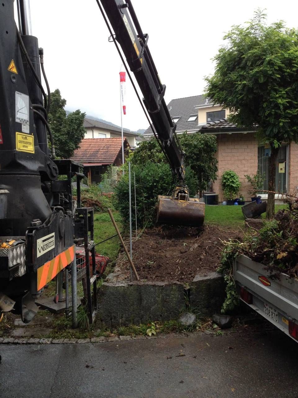 Ein Bagger hebt Erde in einem feuchten Wohngarten neben einem Haus und einem geparkten Anhänger an.