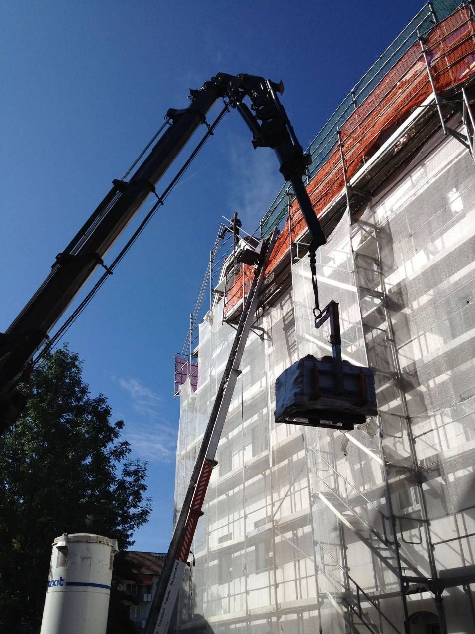 Ein Kran hebt eine Hängeplattform neben einem im Bau befindlichen Gebäude an, das mit Gerüst und Netzen gesichert ist.