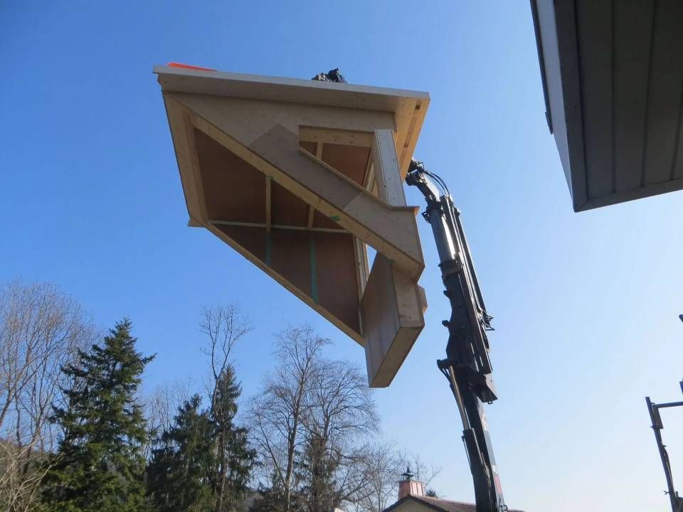 Ein Bagger hebt ein grosses dreieckiges Dachsegment vor blauem Himmel im Freien an.