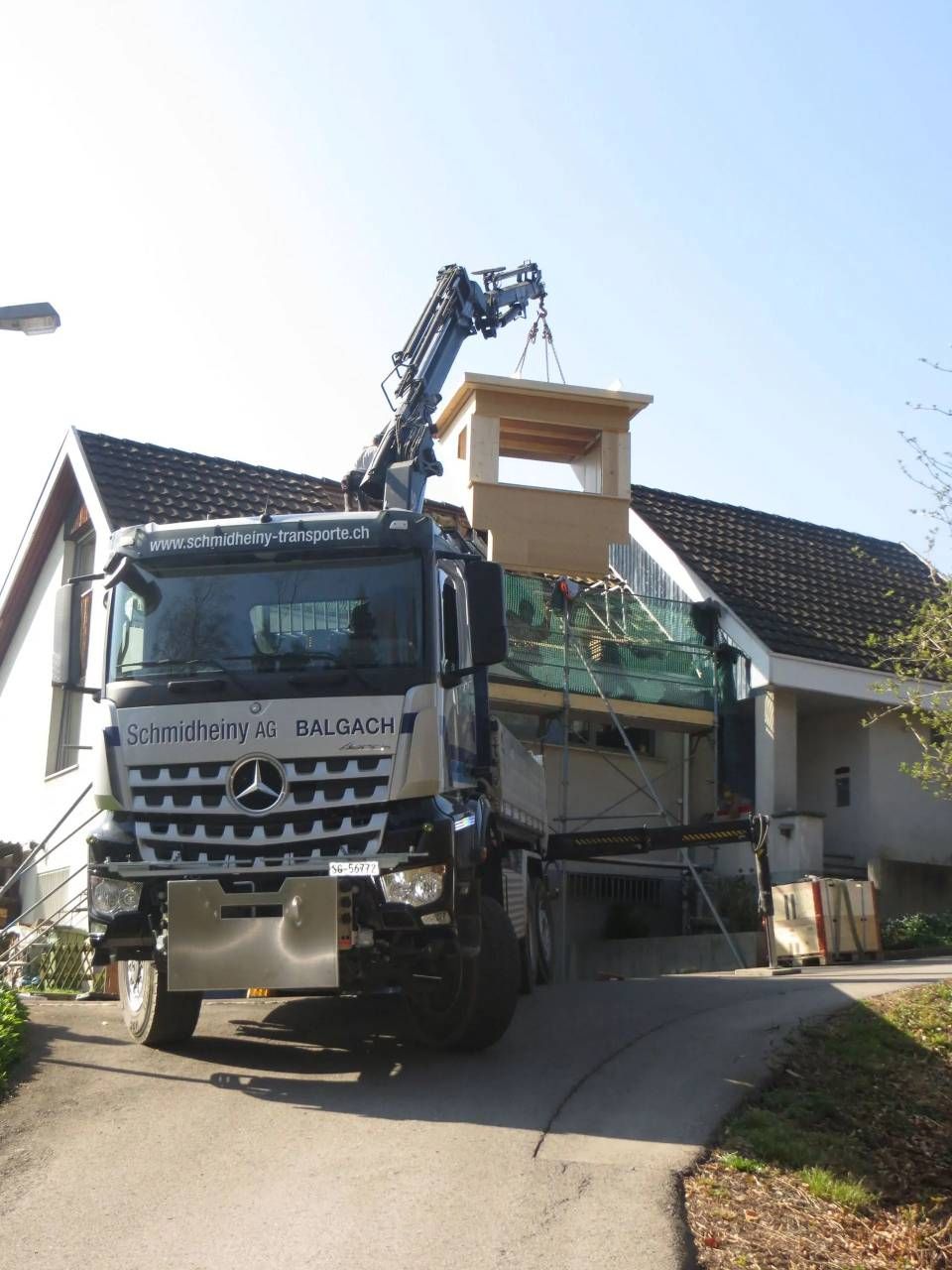 Ein Kranwagen hebt ein kleines Hausteil auf ein Wohngrundstück neben einer steilen Zufahrt.