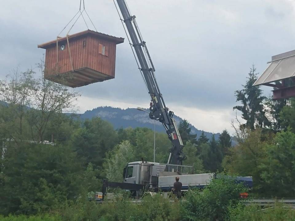 Ein Kran hebt eine kleine Holzhütte über eine ländliche Landschaft mit Bergen und Bäumen.