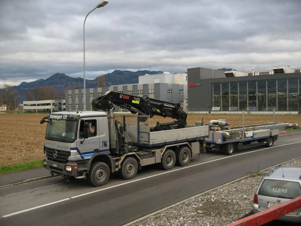 Ein Kranwagen zieht einen Tieflader auf einer Strasse neben einem Industriegebäude.