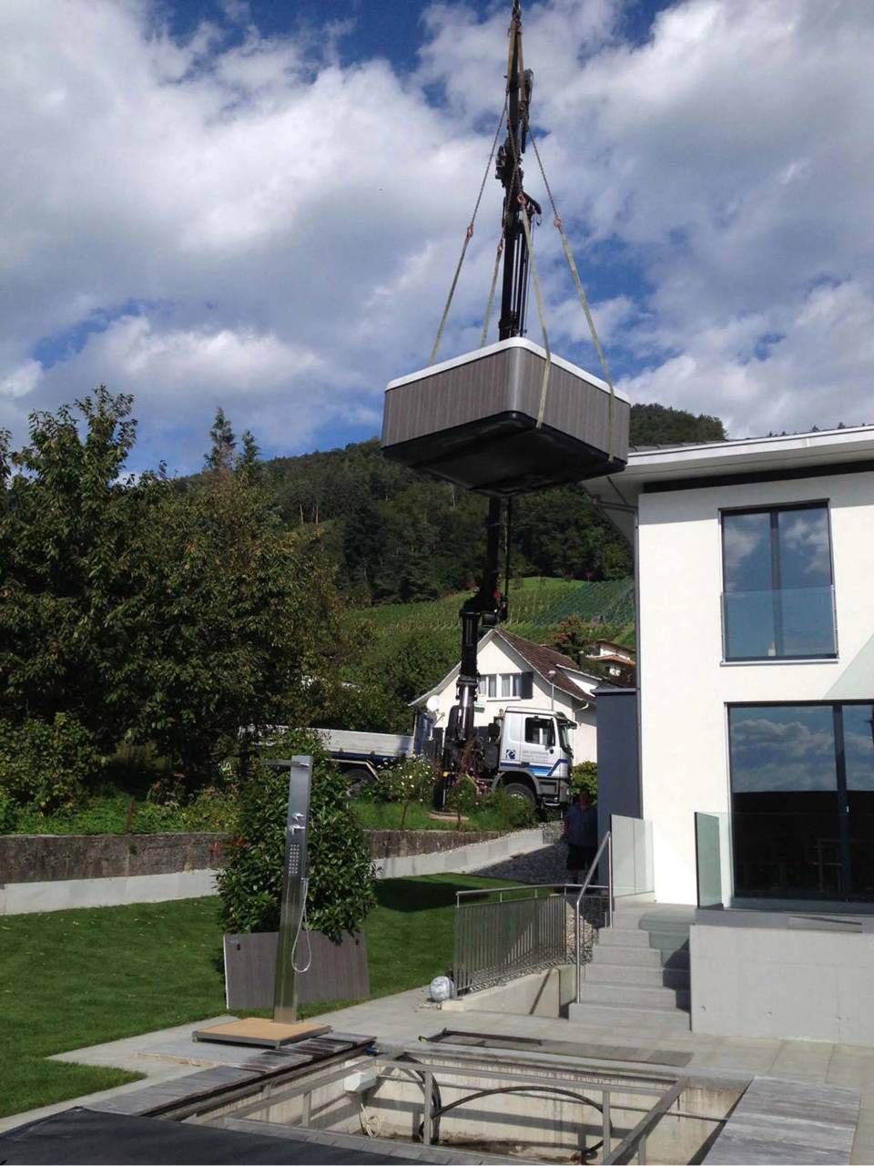 Eine grosse Kiste hängt an einem Kran neben einem modernen Haus; im Hintergrund sind ein bewölkter Himmel und eine Hügellandschaft zu sehen.