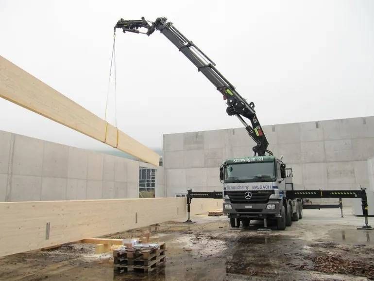 Ein LKW-Kran hebt einen langen Balken auf einer Baustelle mit Betonwänden an.