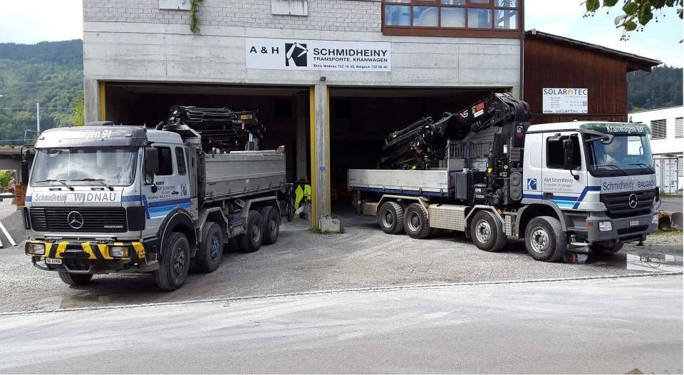 Zwei Muldenkipper parkten vor einer Garage mit offenen Toren und einem Gebäudeschild darüber.