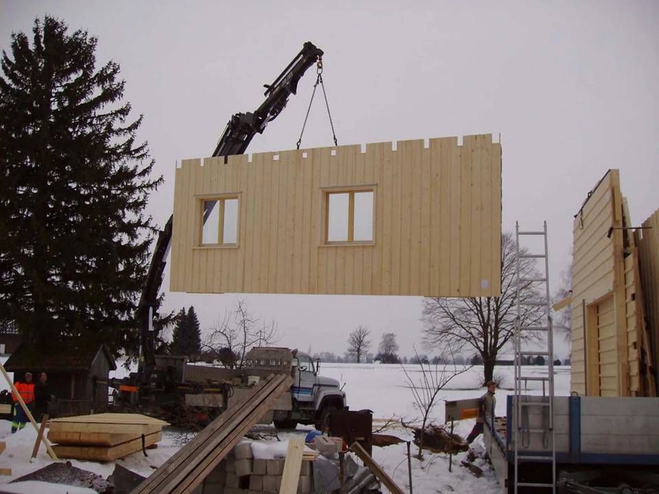 Ein Kran hebt eine grosse hölzerne Wandplatte an einer verschneiten Baustelle an.