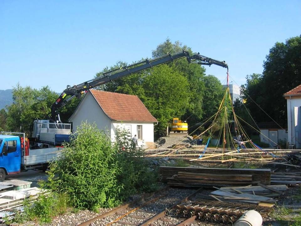 Baustelle mit Kran, der neben weissen Häusern und Bäumen unter blauem Himmel Baumaterialien hebt