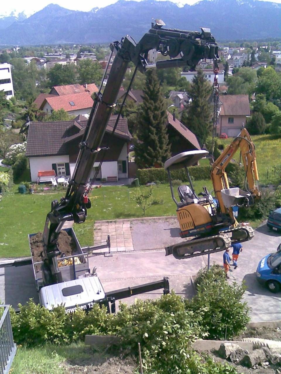 Zwei Bagger heben eine lange schwarze Stange in einer Wohnstrasse an; im Hintergrund sind Berge zu sehen.