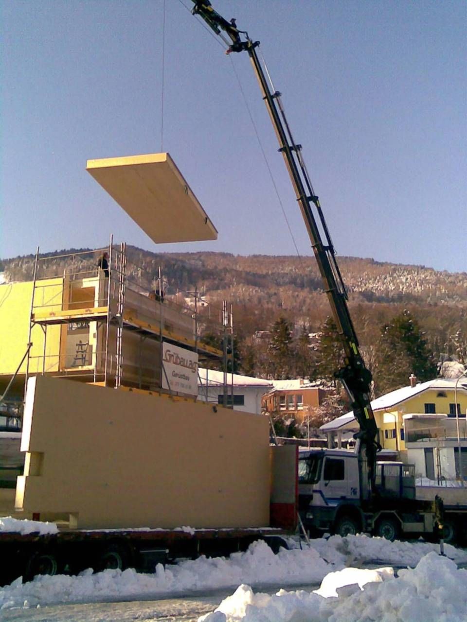 Ein Kran hebt eine grosse Holzplatte auf ein im Bau befindliches Gebäude in einer verschneiten Landschaft.