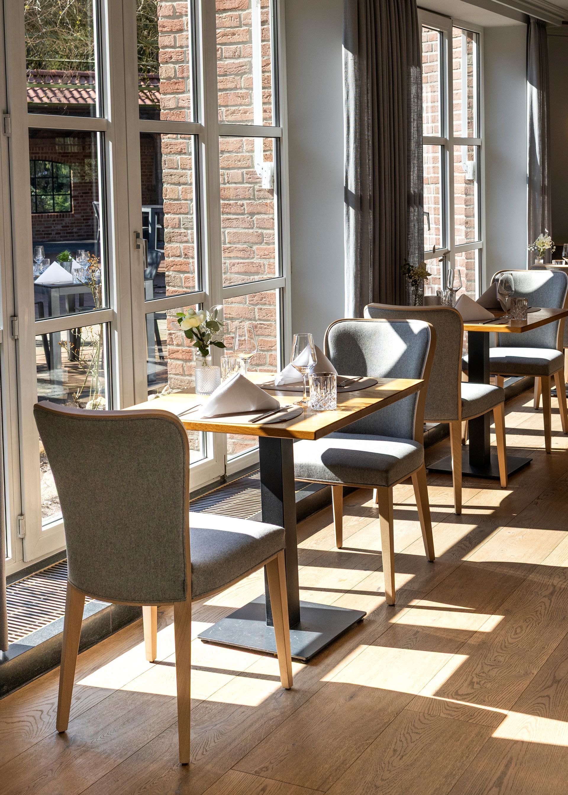 Lichtdurchfluteter Restaurantinnenraum mit gedeckten Tischen, grau gepolsterten Stühlen und hohen Fenstern.
