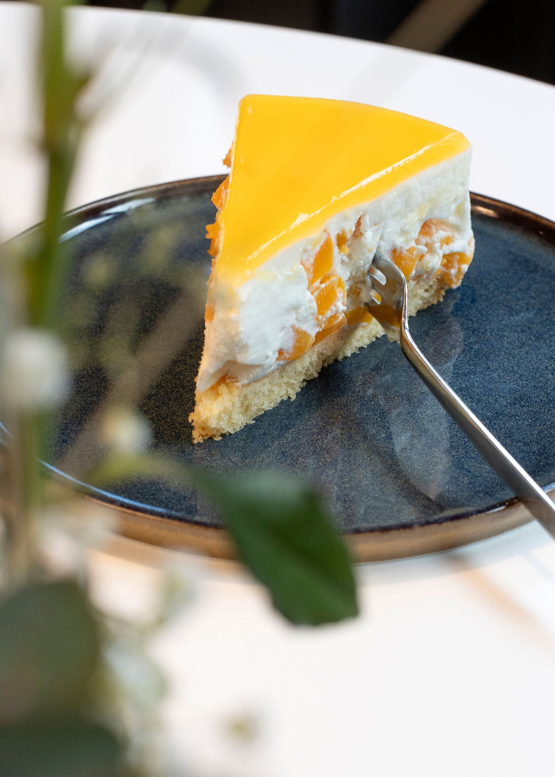 A slice of mango mousse cake with yellow icing and pieces of fruit on a dark blue plate, along with a fork and a blurred plant.