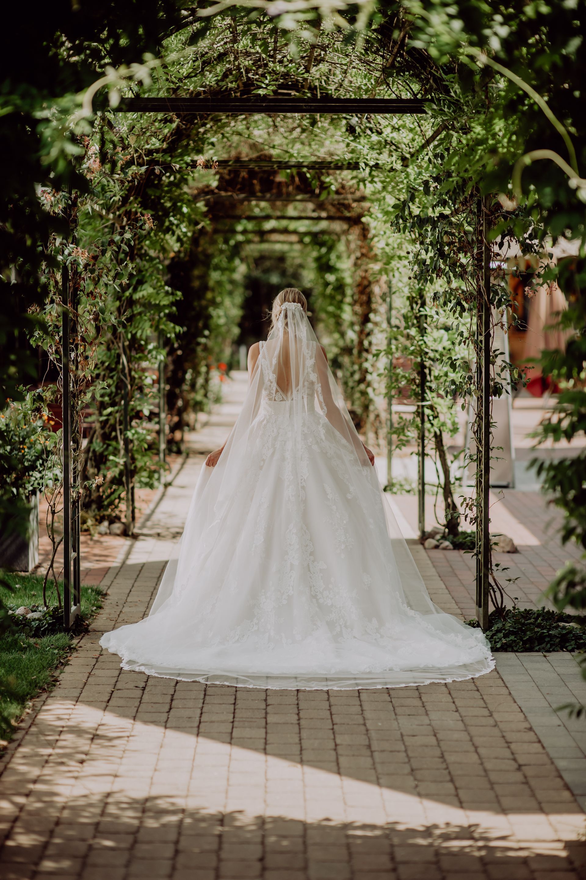 Eine Person in einem weißen Brautkleid mit langer Schleppe schreitet einen gepflasterten Weg entlang, der von einem grünen, bogenförmigen Rankgitter überdacht ist.